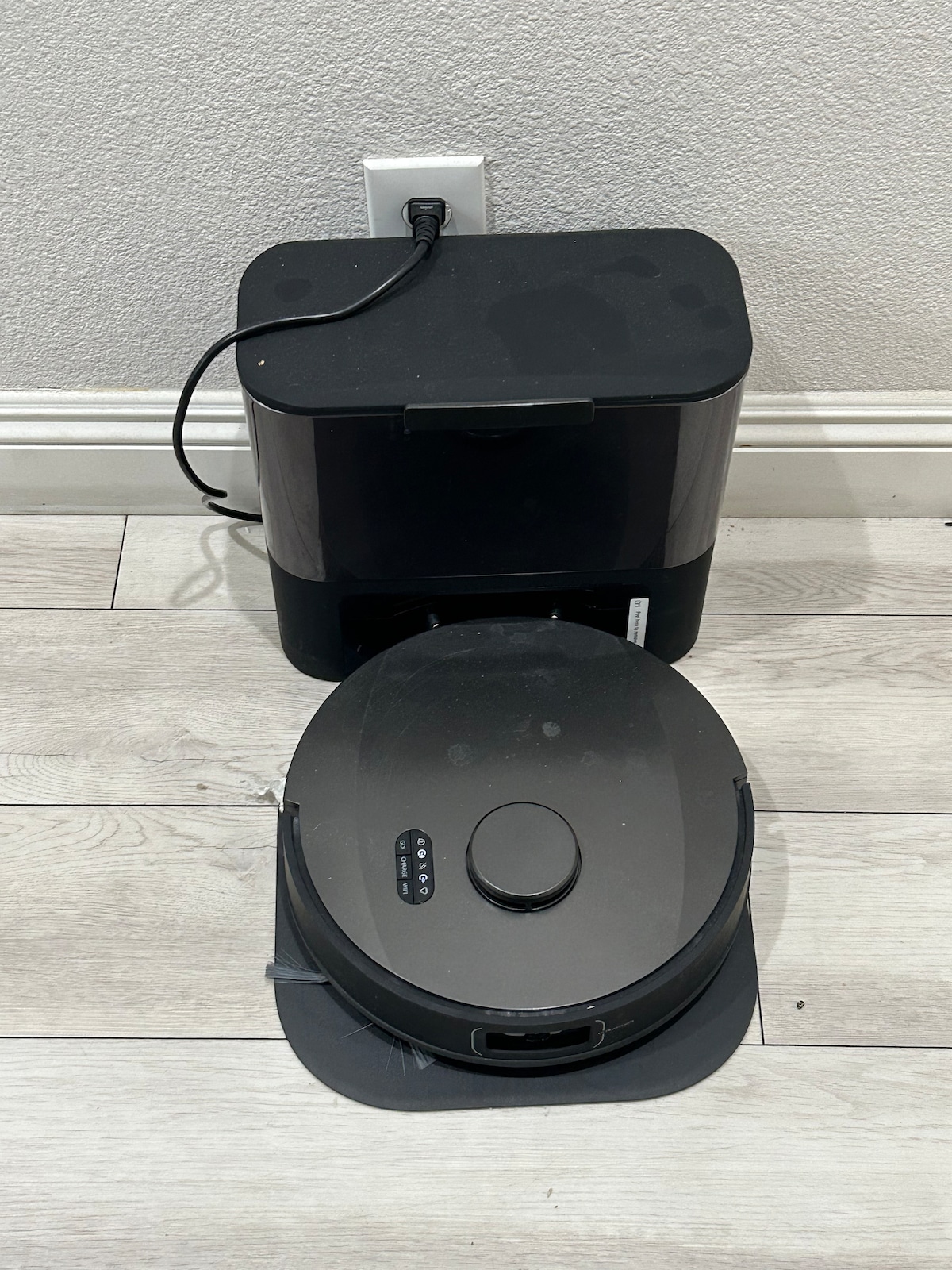 An automated vacuum cleaner is positioned on a light-colored wooden floor. The device is partially docked with its charging station, which is mounted against a light wall. A power cord connects the dock to a wall outlet.