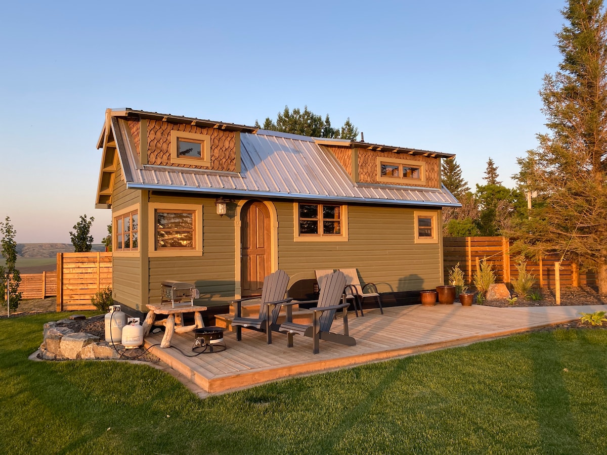 A charming tiny home is set amidst a well-kept yard, featuring a welcoming entrance with an arched door. Outdoor seating is provided by a wooden deck equipped with Adirondack chairs, complemented by surrounding greenery and a fenced area that ensures privacy.