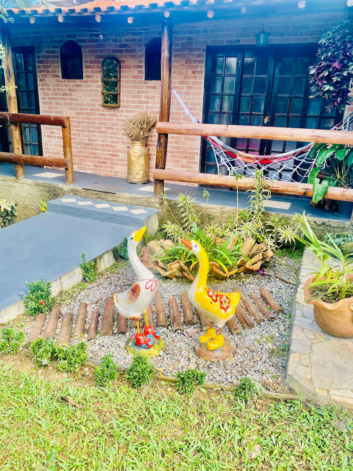 A charming outdoor area is highlighted by colorful decorative ducks positioned among stones and greenery. The rustic brick building with wooden accents is visible in the background, complemented by a hammock and vibrant plants, creating a serene and inviting environment.