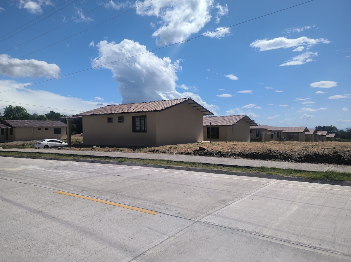 A view of the accommodation showcases the single-story buildings with earthy-toned exteriors. The surrounding area is open, featuring a well-maintained road and grassy patches. The sky is bright, filled with scattered clouds, contributing to a relaxed, natural setting.