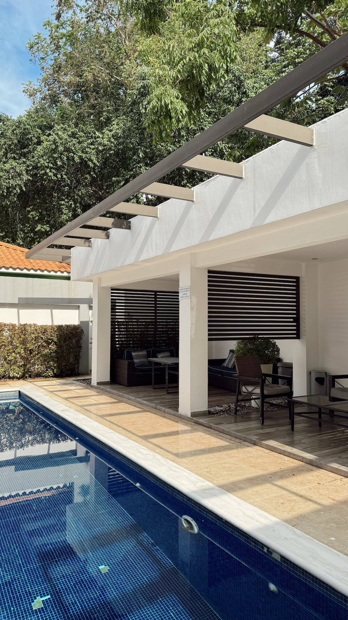A modern outdoor patio area is visible beside a shimmering pool. Shaded seating arrangements under a pergola provide a relaxing space, with lush greenery surrounding the area. The pool reflects sunlight, enhancing the serene ambiance of the outdoor environment.