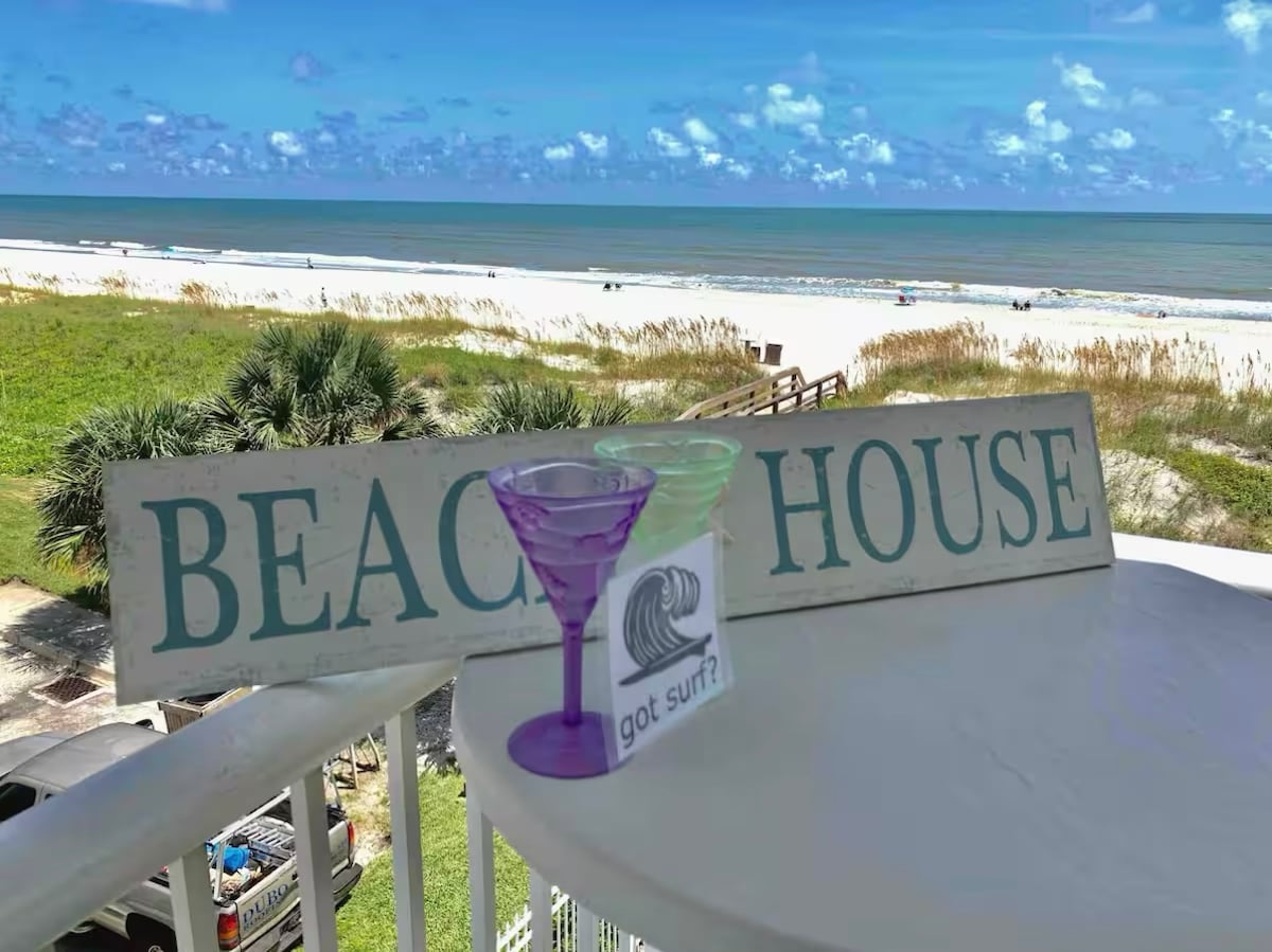 A sign reading 'BEACH HOUSE' is displayed on a table, accompanied by colorful cocktail glasses. The view beyond features a sandy beach and gentle waves under a clear blue sky, with lush greenery in the foreground.