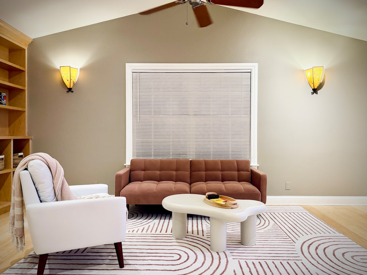 A spacious living area is highlighted by a large window featuring horizontal blinds. A brown sofa and a white accent chair are positioned nearby a rounded coffee table, alongside a patterned area rug that adds texture to the space. Decorative wall sconces provide soft lighting.