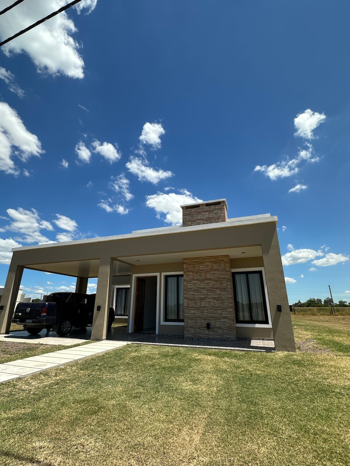 A modern house is presented with a stone accent façade and a covered porch. The structure features large windows, allowing natural light to enter. The front lawn is neatly trimmed and extends out to an open landscape under a bright blue sky with scattered clouds.