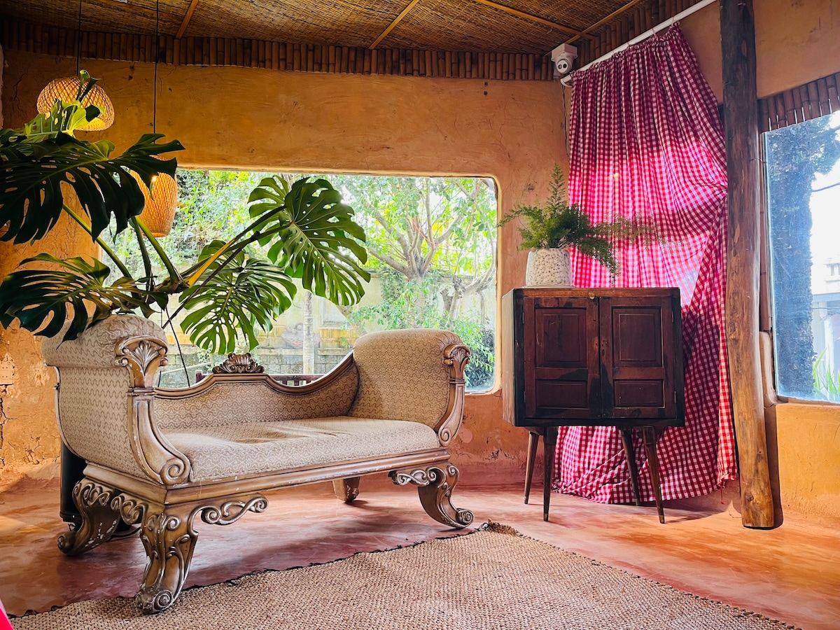 A cozy seating area is highlighted by a vintage-style couch with intricate wooden detailing. Sunlight filters through large windows, illuminating the room's earthy tones. A small cabinet stands nearby, complemented by a potted plant and a patterned curtain that adds a rustic touch.