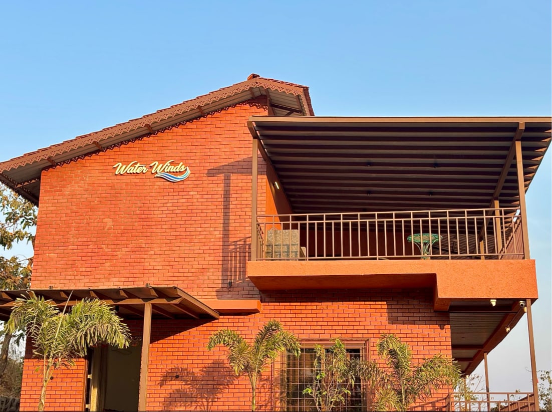 A two-story brick house stands under a clear blue sky, showcasing a balcony with a shaded area. The warm color of the bricks is complemented by the surrounding greenery. The house features a sign that reads 'Water Winds,' adding a charming touch to the exterior.