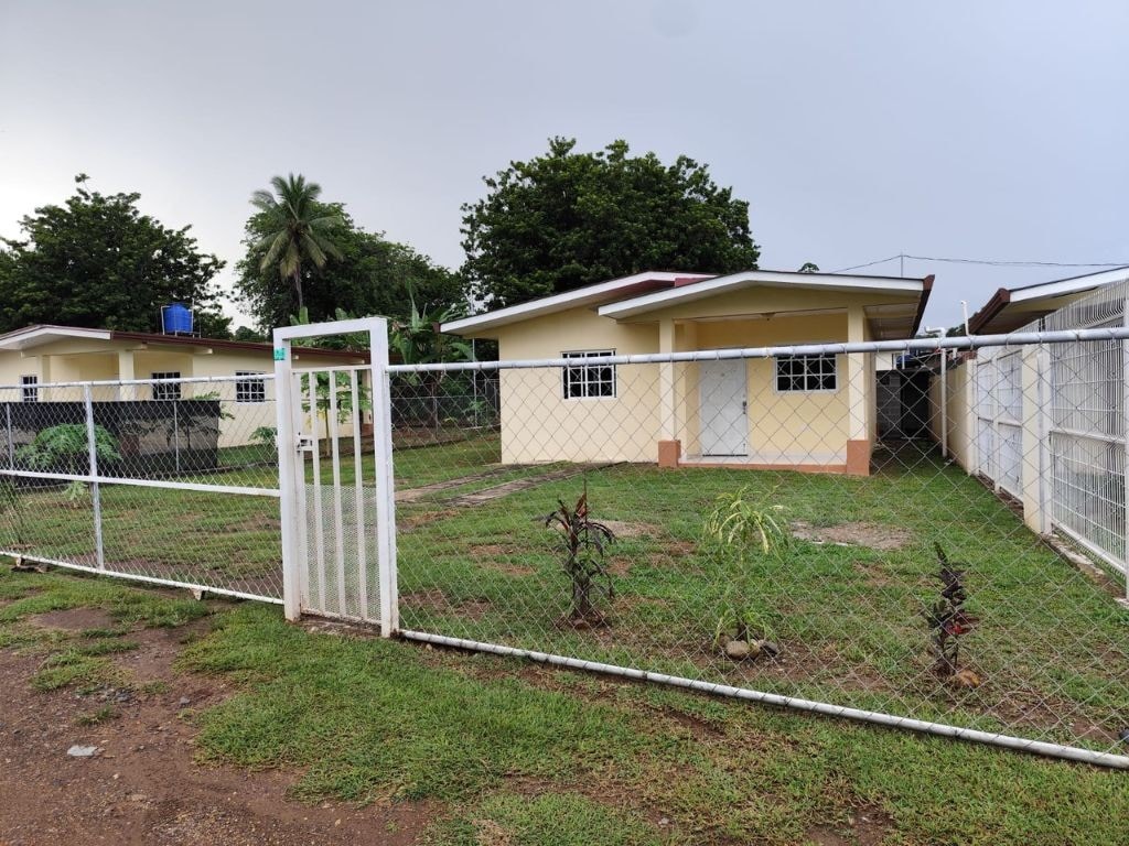 A single-story house is visible behind a fenced yard, featuring a white door and large windows. Lush green plants are present in the foreground, while trees and a water tank are seen in the background, contributing to the serene outdoor environment.