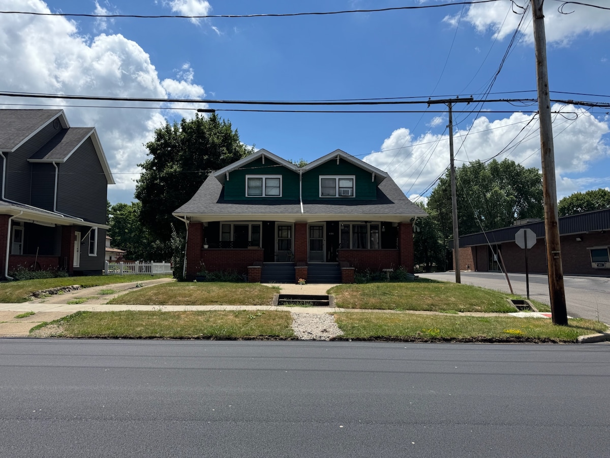 The exterior of a duplex is presented, featuring a symmetrical design with a green upper facade and red brick lower walls. Lush green grass borders the walkway leading to the entrance. Nearby, utility poles and a stop sign are visible along the road.