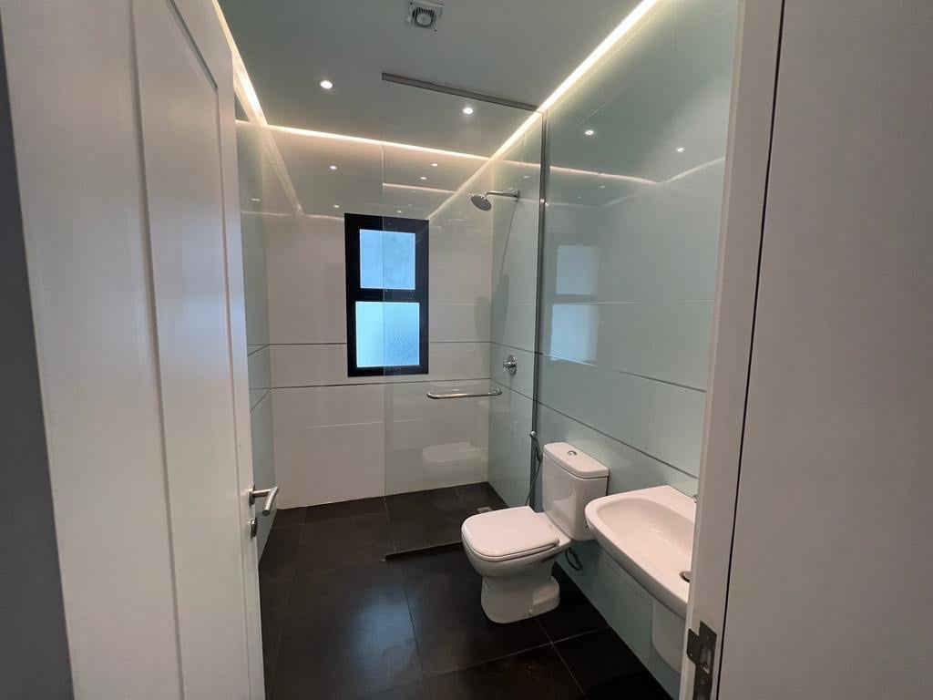 A modern bathroom features sleek white tiles and contemporary fixtures. A glass shower enclosure offers a clear view of the showerhead, while a toilet and a bidet are positioned against the wall. Recessed lighting enhances the clean and spacious feel.