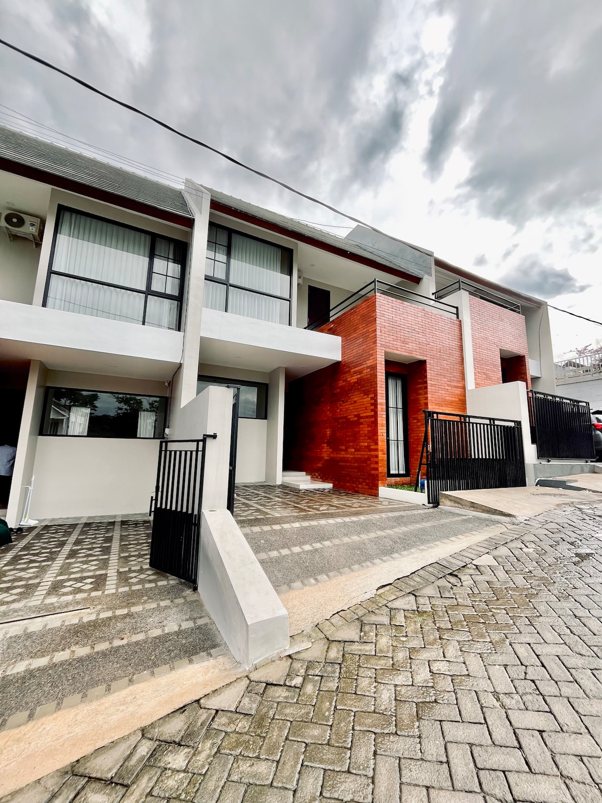A modern, two-story building features a mix of sleek white and warm red brick facades. The entrance is framed by tall glass doors and bordered by black fencing. A paved walkway leads up to the property, creating a tidy and structured appearance.