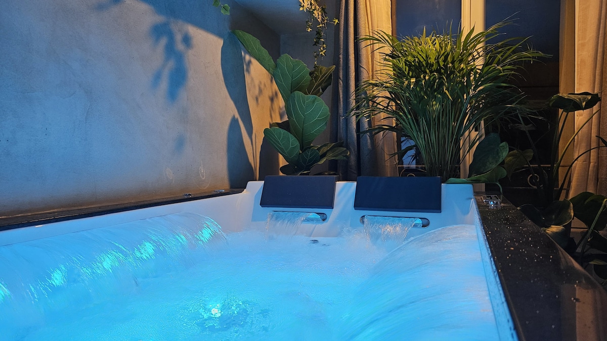 A luxurious hydromassage bathtub is highlighted, featuring 68 jets and illuminated by soft blue lighting. Surrounding plants add a touch of nature, enhancing the serene ambiance. Soft cascading water can be seen flowing, creating a relaxing atmosphere.