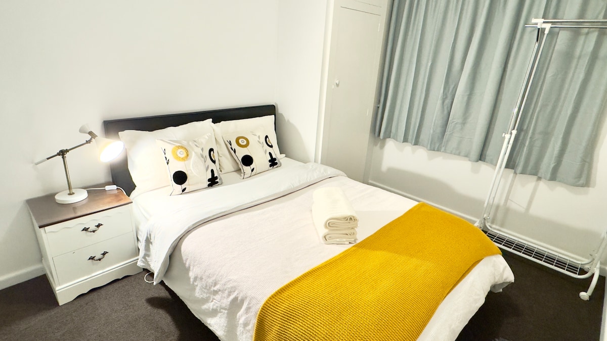 A cozy bedroom features a spacious bed dressed in neutral linens and accented with decorative pillows. A bedside lamp illuminates a side table. Soft light filters through the window, covered by light, flowing curtains. A clothing rack stands nearby, providing additional storage options.