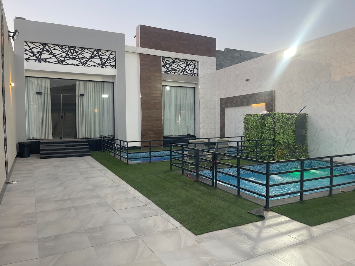An outdoor space features a smooth tiled patio with two small pools surrounded by green grass. The modern structure's large glass doors provide open access, while the patio is bordered by black railings. Soft lighting highlights the adjoining walls, creating a serene environment.