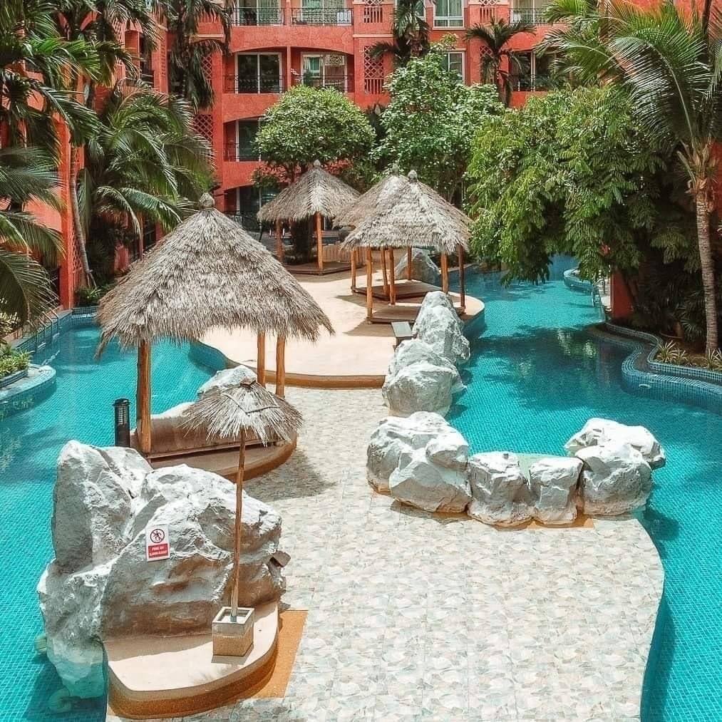 A serene pool area is surrounded by tropical palm trees and lush greenery. Thatched-roof cabanas are positioned on wooden platforms over water, with smooth stones dotting the pathway, creating a tranquil oasis for relaxation.