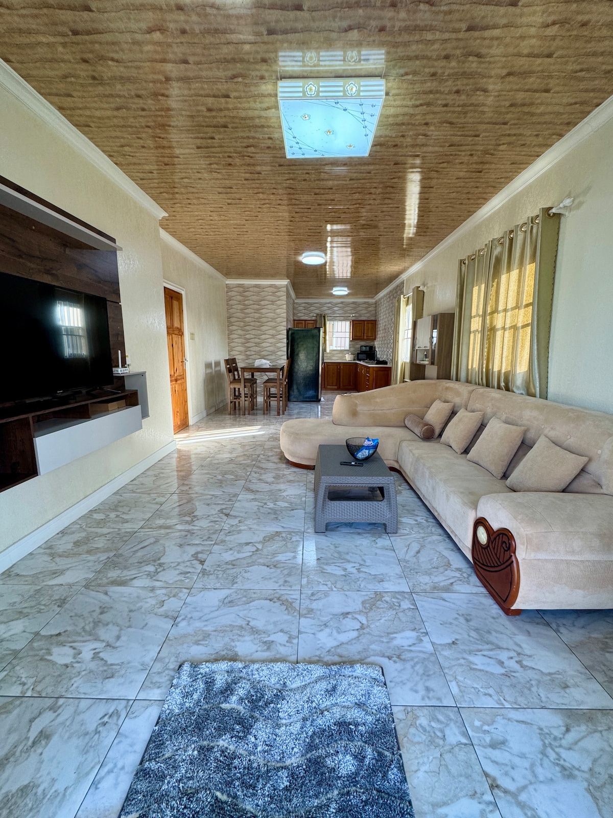 A spacious living area features a light-colored sectional sofa, positioned near a large wall-mounted television. Natural light floods the room through multiple windows, which are framed by soft curtains. A patterned area rug lies beneath a low coffee table, enhancing the inviting ambiance.
