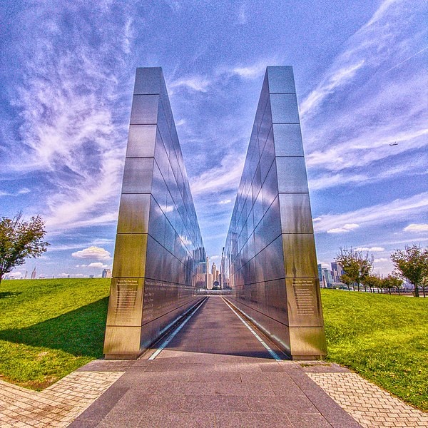 A modern memorial structure is depicted, featuring tall, reflective metal panels that create a corridor-like pathway. Lush greenery on either side enhances the view toward the distant skyline, where urban buildings rise under a blue sky adorned with wispy clouds.