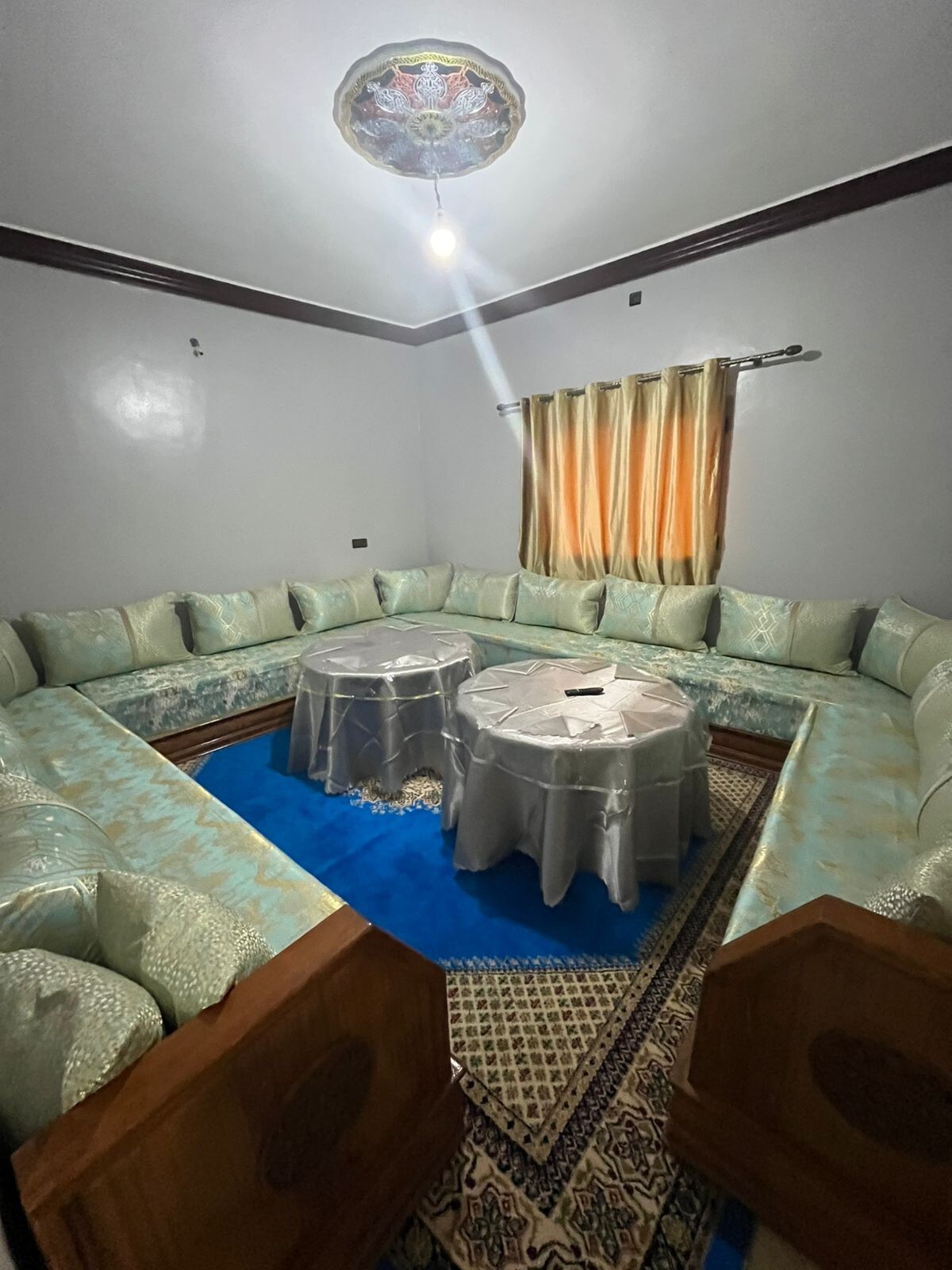 A circular seating arrangement is highlighted by plush, patterned cushions in soft turquoise hues. Two round tables are placed at the center, topped with a tablecloth. Decorative curtains frame the window, allowing warm light to enhance the serene atmosphere.