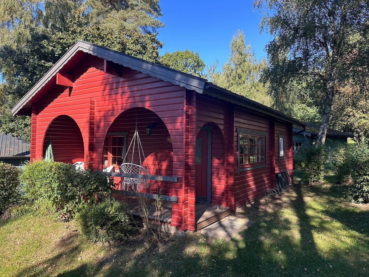 A charming red wooden cottage features arched entryways and a welcoming porch. Surrounded by lush greenery, sunlight casts dappled shadows on the landscape, enhancing the cottage's rustic appeal. Multiple windows provide natural light, inviting guests to enjoy the peaceful outdoor setting.