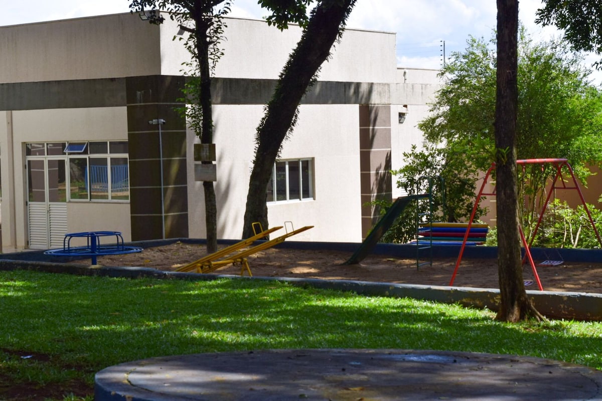 An outdoor play area is visible, featuring colorful swings and a yellow see-saw set on a soft grassy surface. The building provides a neutral backdrop with large windows, allowing natural light to enter. Surrounding trees offer shade and a sense of tranquility.