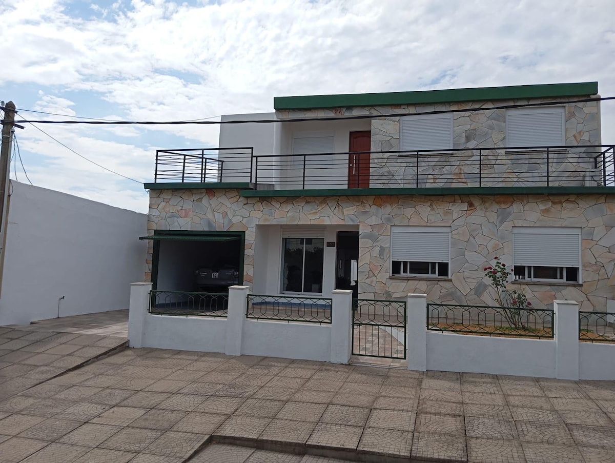 A two-story house features a stone exterior and a green roof. The front is accessible via a gated walkway, with large windows on both levels and a garage space visible. A small garden with plants is located along the front facade.