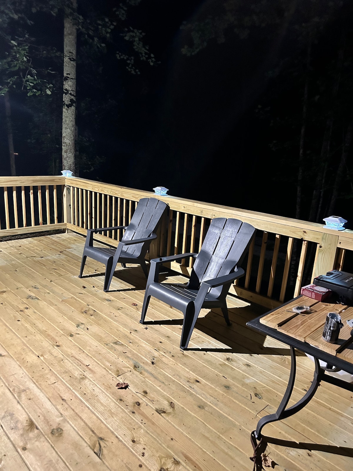 A wooden deck at night is featured, illuminated by subtle lighting. Two dark-colored adirondack chairs are positioned for relaxation, accompanied by a small table that holds various items. The surrounding area includes trees, creating a secluded atmosphere.