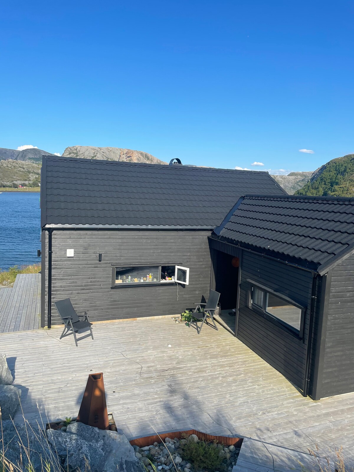 A modern black cabin is situated near a tranquil body of water, with mountains visible in the background. A wooden deck is featured in front of the cabin, equipped with a few outdoor chairs. Large windows enhance natural light inside the structure.