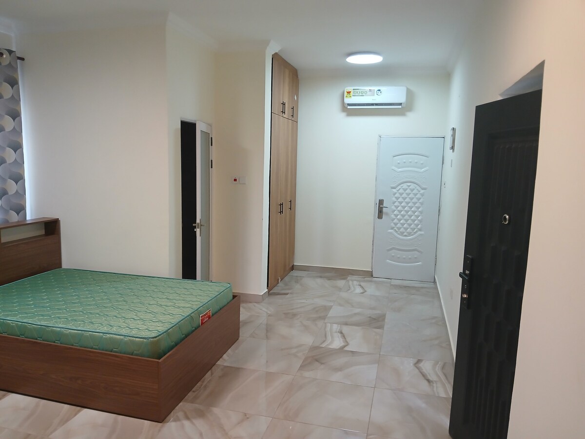 A well-lit room features a spacious bed with a green mattress and a large wardrobe for storage. An air conditioning unit is mounted on the wall, while the entrance door stands at the far end of the room. Neutral-colored walls and tiled flooring enhance the space.