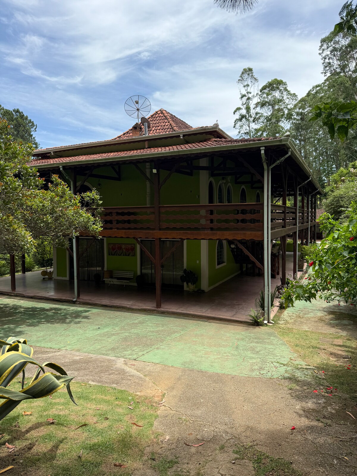 The green exterior of a spacious, two-story house is framed by lush trees, showcasing a porch with wooden railings and a terra-cotta tiled roof. The ground is partially paved, with a pathway leading towards the front door. An outdoor satellite dish is visible on the roof.