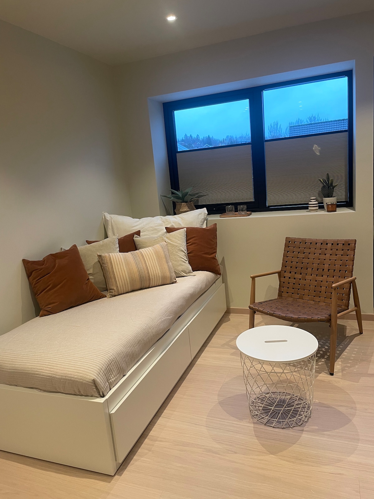 A cozy living area features a light-colored daybed adorned with a variety of pillows. A simple wooden chair and a round coffee table in front create a welcoming space. Large windows allow natural light to filter in, enhancing the calming atmosphere.