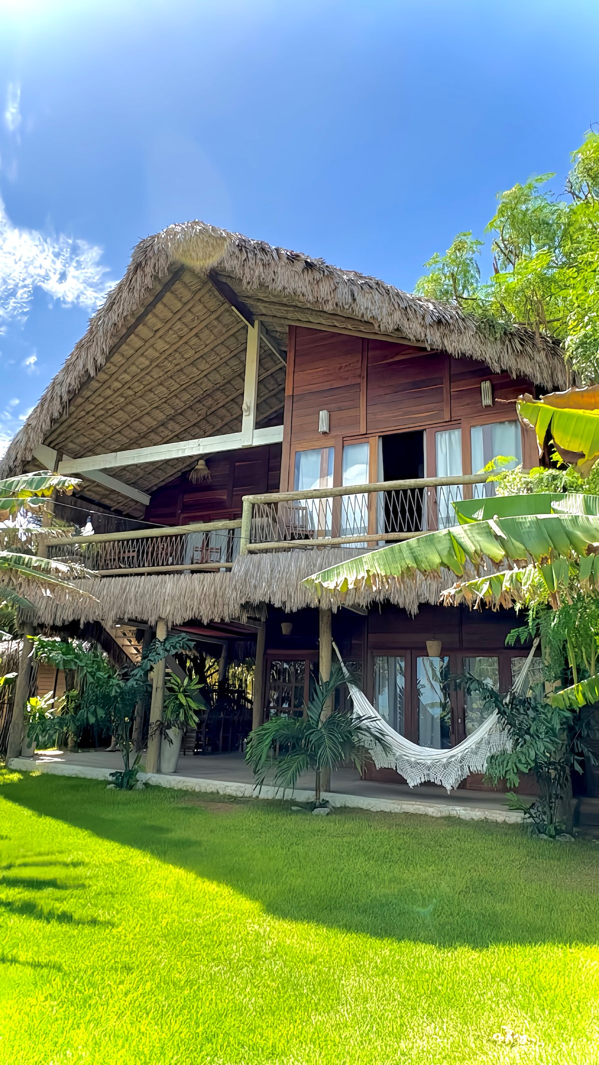 A charming two-story bungalow is depicted, featuring a thatched roof and wooden accents. Lush greenery surrounds the structure, while a hammock is suspended in the shaded area below. Large windows offer ample natural light, contributing to a warm and inviting appearance.