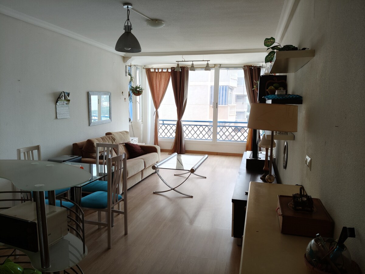A bright living area is shown, featuring a sofa with soft pillows and a glass coffee table. A dining table with chairs is positioned nearby. Large windows allow natural light to fill the space, with light curtains enhancing the airy atmosphere.