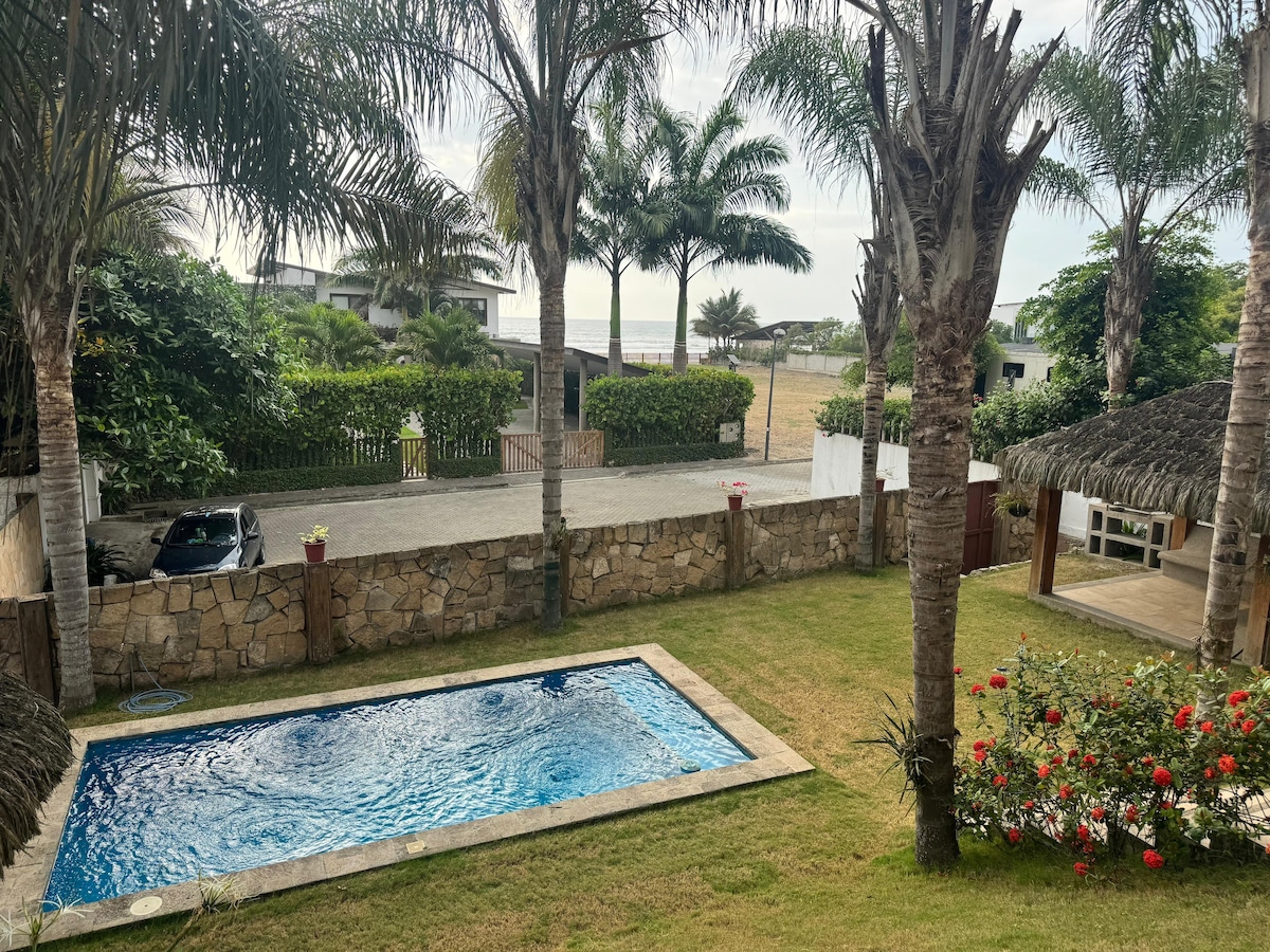 A sparkling pool is framed by a stone patio, surrounded by lush greenery and palm trees. The vibrant garden features blooming flowers, while the peaceful landscape leads to the ocean in the distance, creating a serene outdoor space perfect for relaxation.