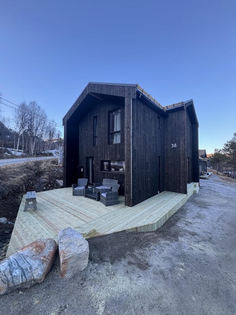 The modern cabin exterior features dark wooden paneling and a unique architectural design. A spacious wooden deck is adorned with two seating chairs, providing an inviting space for relaxation. The surroundings are characterized by natural elements, with gravel pathways and hillside trees in the background.