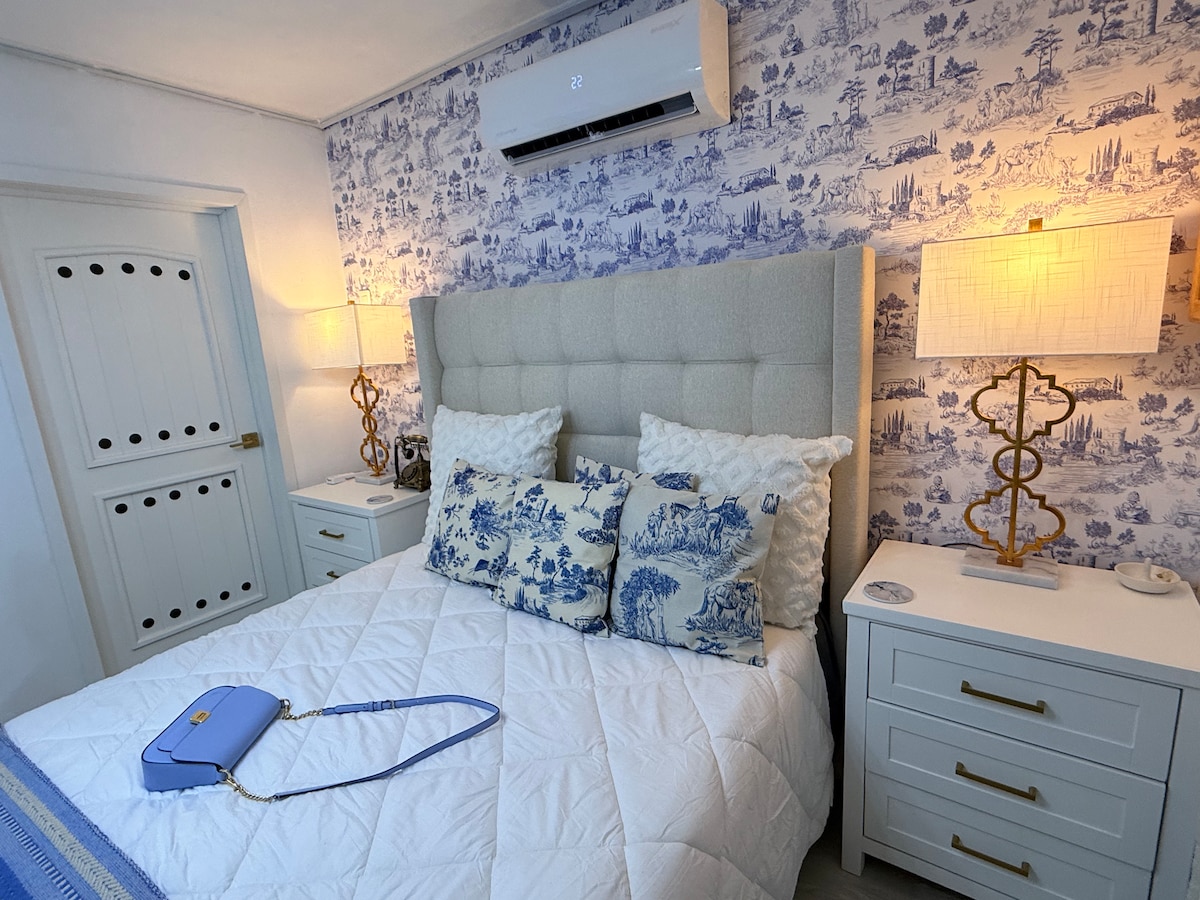 A cozy bedroom features a queen bed with a light-colored upholstered headboard. Decorative blue and white pillows are arranged on the bed. Two lamps beside white nightstands provide soft lighting, while a stylish wallpaper adds a charming backdrop.
