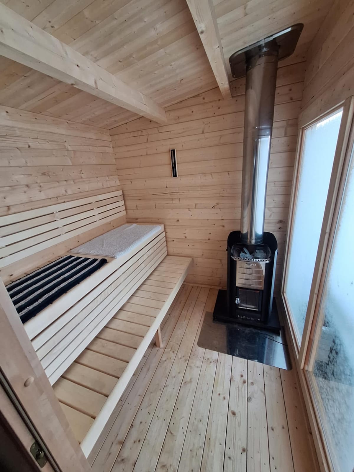 The interior of a sauna features warm wooden paneling with benches along the walls. A stove is positioned in the corner, emitting heat. Natural light filters through a frosted glass window, illuminating the serene space.