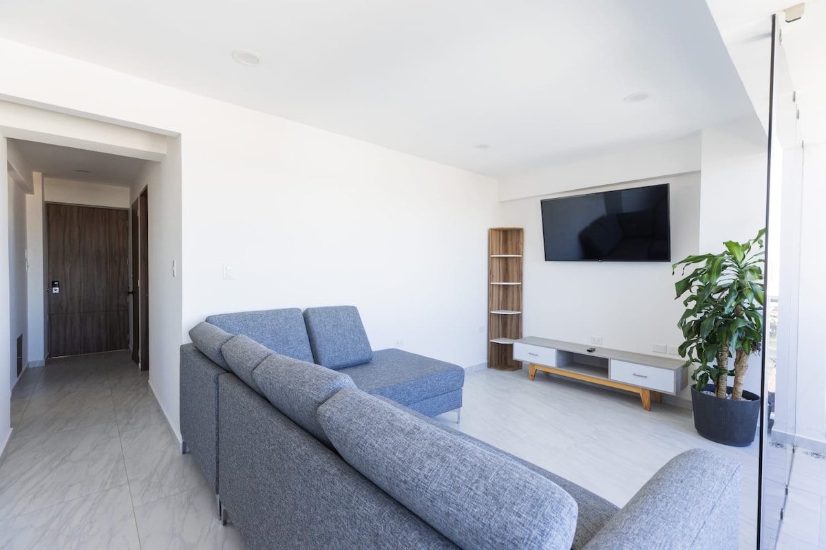 A bright living area features a spacious gray sectional sofa positioned near a wall-mounted television. A sleek entertainment unit is below the TV, complemented by a tall bookshelf. Natural light fills the space through large windows, enhancing the minimalist design.