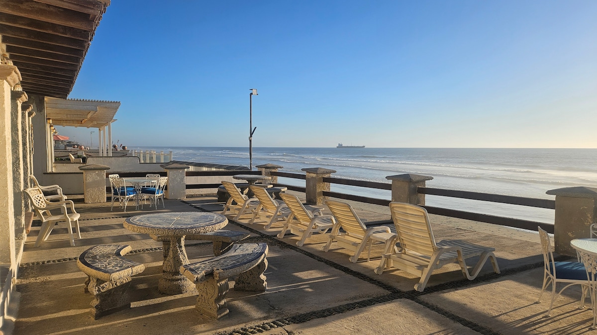A spacious beachfront patio is captured under a clear blue sky, showcasing multiple cushioned lounge chairs and stone-tiled tables. The calm ocean glistens in the sunlight, with a distant ship visible on the horizon. The railing adds a picturesque boundary to the tranquil seaside view.