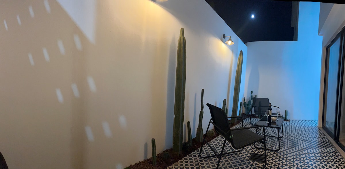 A serene outdoor space is presented with two black lounge chairs set on a patterned floor. Tall cacti are painted on the wall, complemented by soft lighting that highlights their silhouettes against the backdrop of a dark sky, featuring a visible moon.