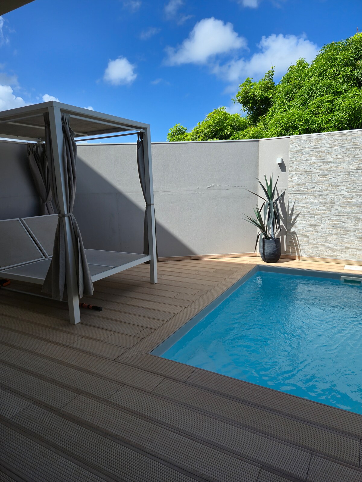 A private pool area features a clear blue pool framed by wooden decking. A comfortable outdoor lounge with draped privacy curtains and a potted plant is positioned alongside the pool, providing a serene retreat surrounded by greenery.