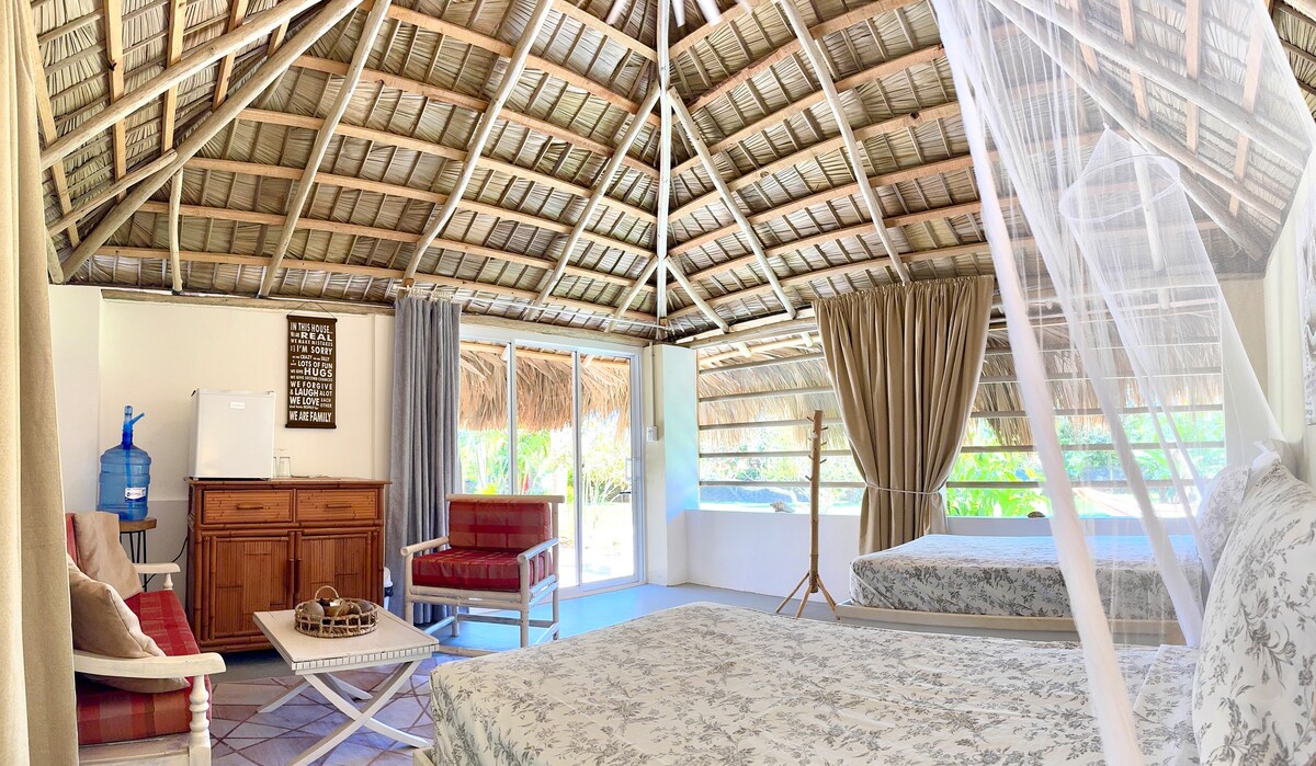 An open-concept space showcases a high thatched roof, with large windows allowing natural light to fill the room. Two spacious beds are positioned alongside a cozy seating area that includes a wooden table and chairs. A water dispenser is visible near a wooden cabinet.