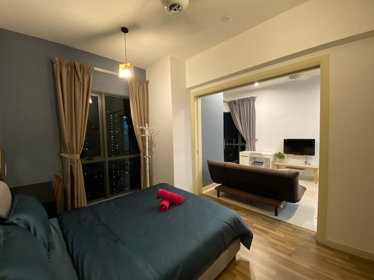 A comfortable bedroom is presented with a spacious bed featuring dark blue bedding and a rolled towel. Natural light enters through large windows, framed by contemporary curtains. A door leads to a living area, which includes a sofa and a television.