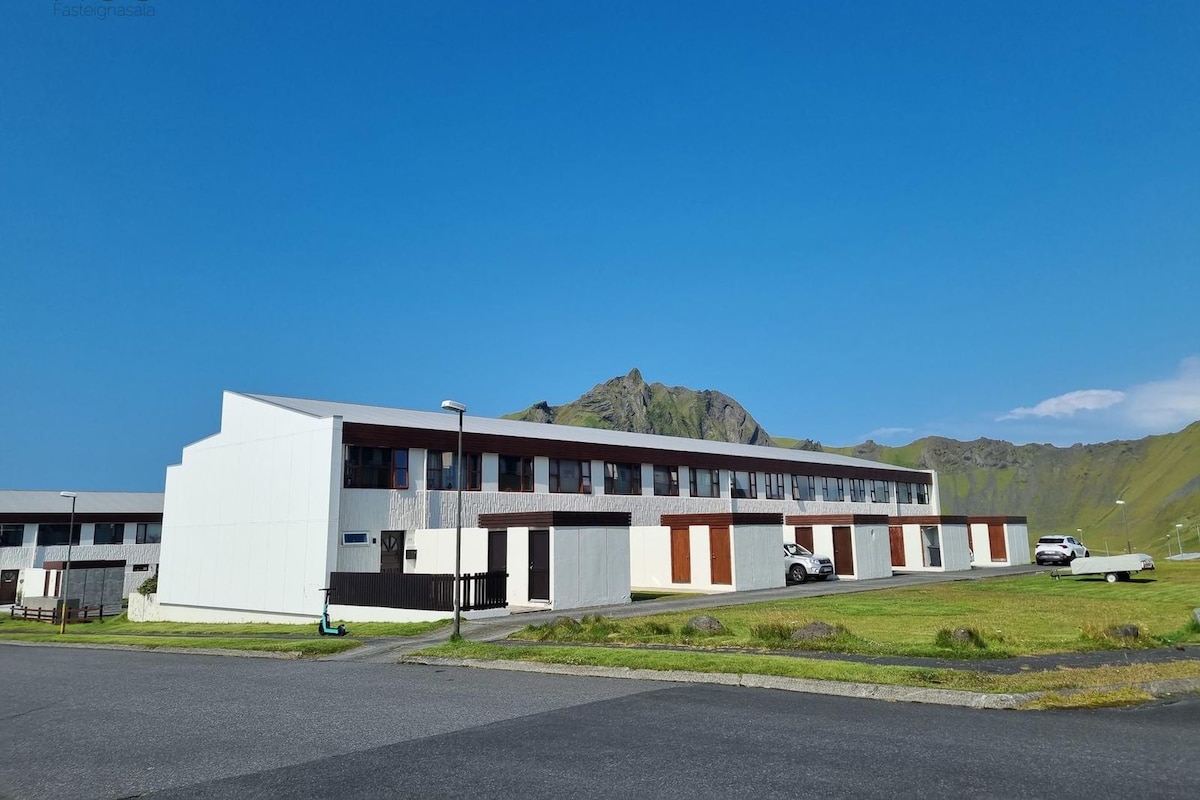The apartment building is displayed, showcasing a modern architectural design with multiple windows and a mix of wooden and white exterior. The backdrop features a green hillside under a clear blue sky, providing a serene setting.
