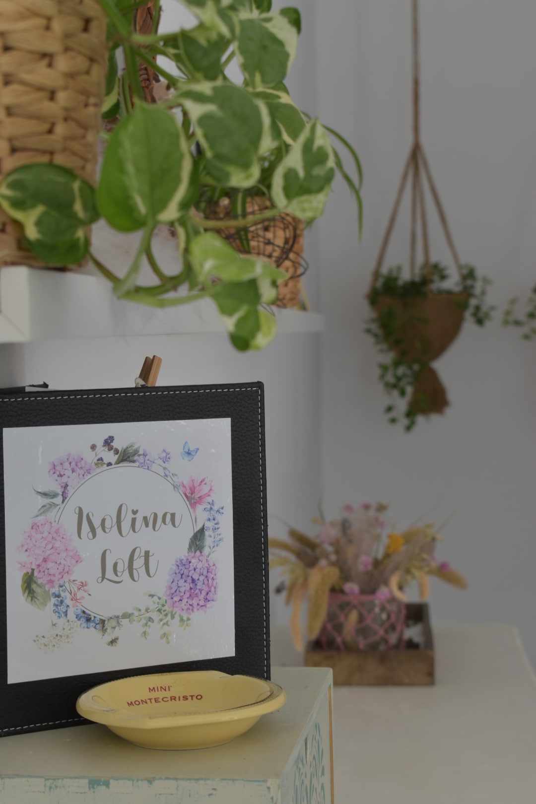 A framed sign titled 'Isolina Loft' decorated with floral designs is positioned on a light-colored surface. Lush green plants are incorporated into the decor, with a hanging planter visible in the background, enhancing the natural ambiance of the space.