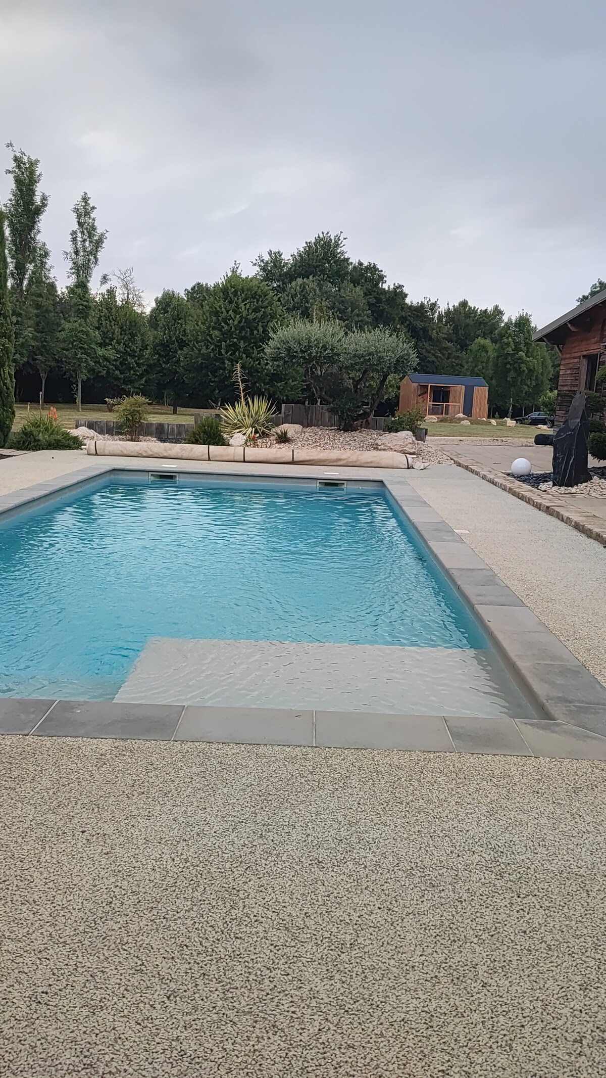 A rectangular pool is visible, surrounded by a pebble walkway and landscaped gardens. Lush greenery and trees are positioned in the background. A small structure is also depicted beyond the pool area, complementing the natural setting.