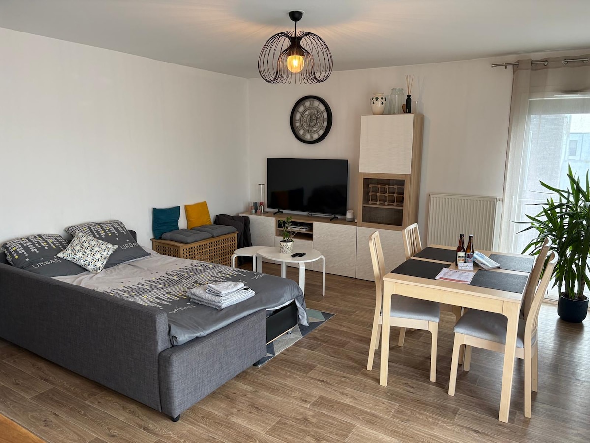 A modern living area features a comfortable sofa bed, a dining table set for four, and a flat-screen TV mounted on a light-colored cabinet. Natural light enters through a window dressed with sheer curtains, complemented by plants enhancing the space's freshness.