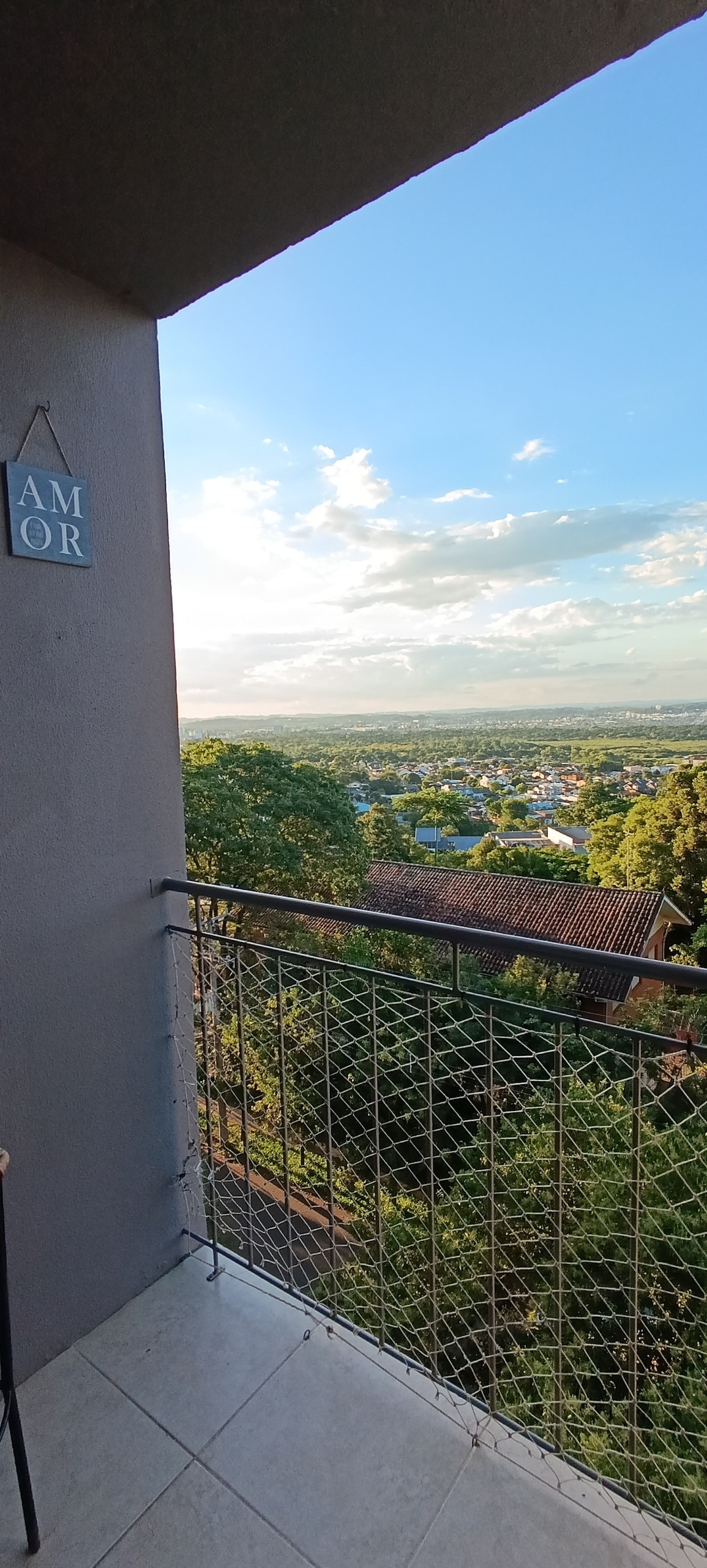 A balcony is seen overlooking a scenic view, featuring green treetops and residential rooftops. The sky reflects a soft blue with scattered clouds, indicating a pleasant day. A decorative sign hangs on the wall, providing an inviting touch to the outdoor space.