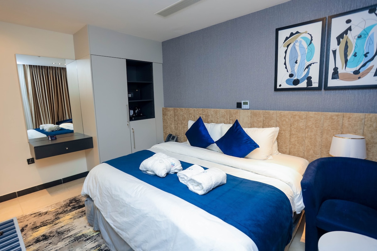 A modern bedroom features a spacious bed adorned with white and deep blue bedding. Two blue accent pillows are placed on the bed. A stylish chair and a large mirror are visible, along with artwork on the wall, enhancing the room's contemporary feel.