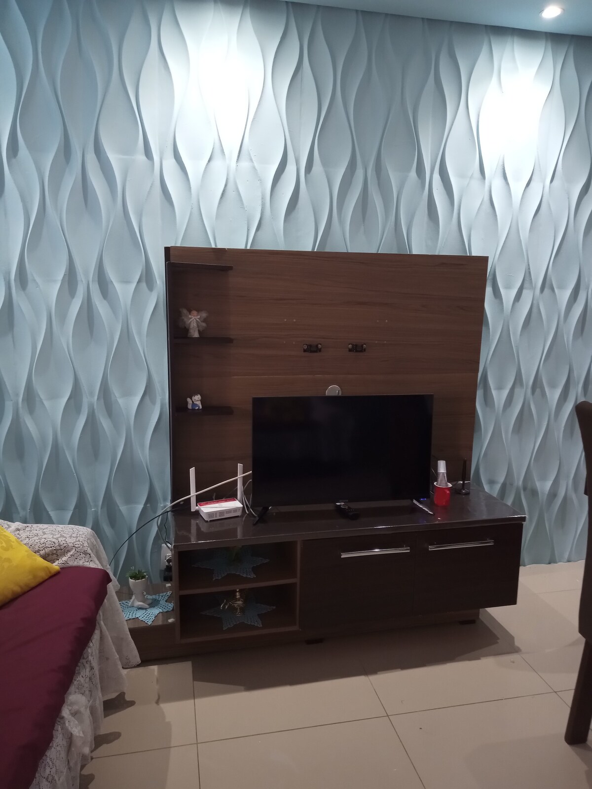 A stylish living area is presented, featuring a wooden entertainment unit with a mounted television and decorative shelves. A textured blue wall adds depth, while a cozy seating area is defined by a soft blanket draped over the side of a bed. Light tiles enhance the room’s brightness.