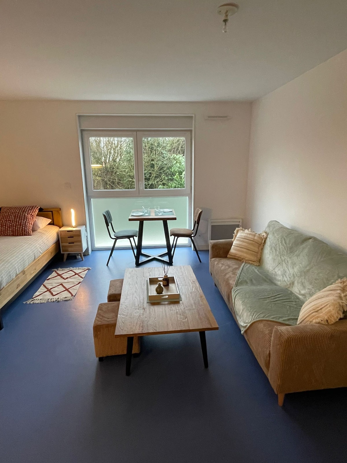 The studio is equipped with a spacious bed on one side and a cozy sofa on the other. A wooden coffee table is placed between the sofa and a dining table set for two. Large windows bring natural light into the room, complemented by neutral decor.