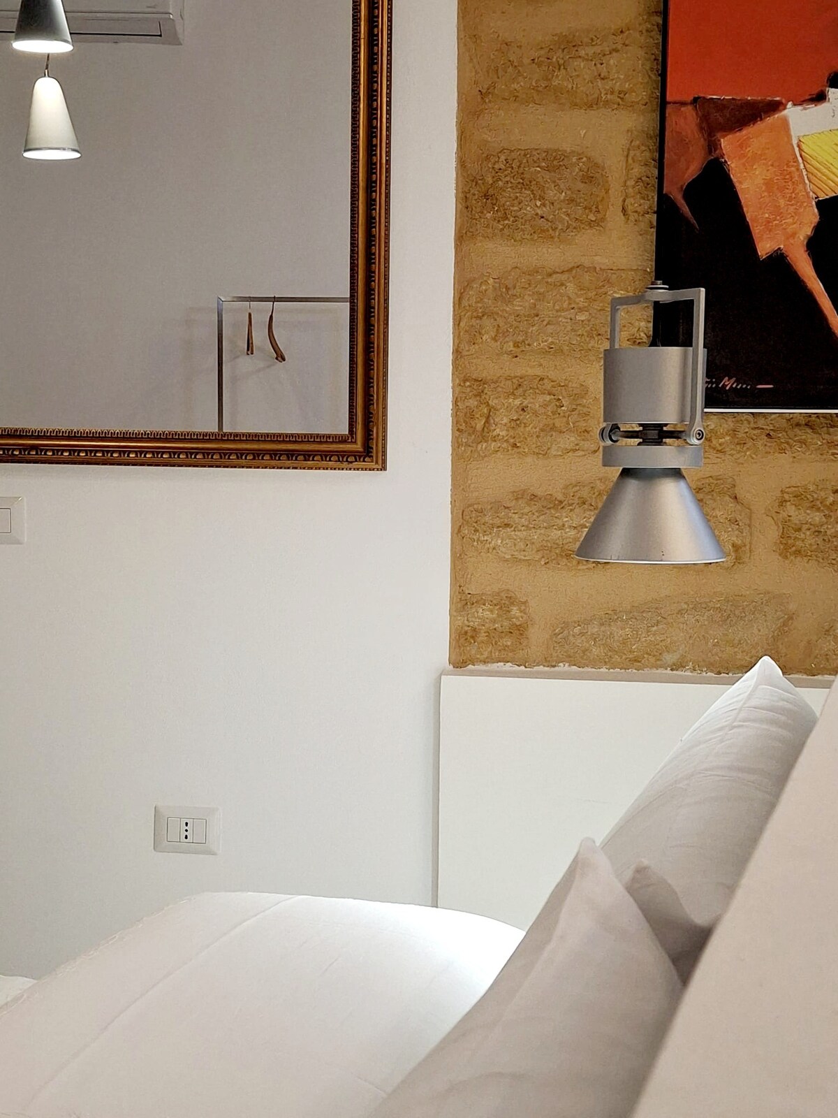A close-up of a cozy sleeping area showcases soft white bedding with pillows positioned against a light wall. A sleek silver lamp hangs beside a modern piece of artwork, while a decorative mirror reflects the surroundings.
